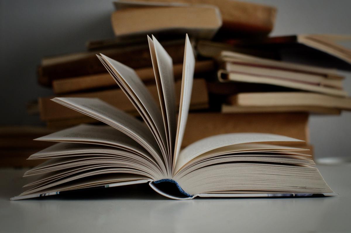 Close up of a hardcover book open with the pages fanned and a few messy piles of books in the background.